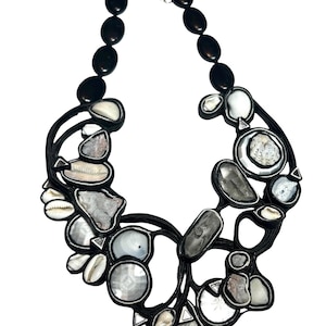 Peut inclure: Un collier original avec un design en cordon noir, intégrant des éléments naturels. Il présente des perles ovales noires, des coquillages blancs et des pierres grises et blanches de forme irrégulière. Un accessoire unique et artistique.