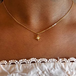 Op de afbeelding: Een delicate gouden ketting met een stervormige hanger met een centrale edelsteen. De ketting wordt getoond tegen een huidtint achtergrond, met een witte kanten rand aan de onderkant. De ketting is fijn en elegant.