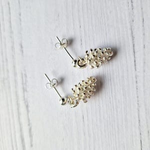 Puede incluir: Un par de pendientes de plata con un diseño de piña. Los pendientes son de estilo colgante con una pequeña bola de plata en la parte superior.