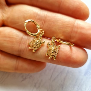 Puede incluir: Un par de pendientes de aro dorados con colgantes de tortuga. Los pendientes presentan colgantes detallados de tortuga colgando de pequeños aros con bisagras. Las tortugas tienen caparazones texturizados y pequeñas patas y cabezas.