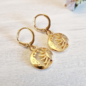 Op de afbeelding: Een paar goudkleurige oorringen. Elke oorring heeft een cirkelvormige bedel met een zon- en maanontwerp. De oorbellen zijn gemaakt van glanzend metaal en worden gepresenteerd op een lichtgekleurd oppervlak.