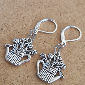 May include: A pair of silver earrings featuring a watering can charm with a floral design. The watering can is filled with flowers and has a heart-shaped handle.