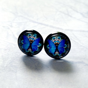 Op de afbeelding: Een paar blauwe vlinder oorbel studs met een zwarte achterkant. De vlinders zijn geplaatst tegen een kleurrijk mandala patroon.