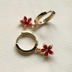 Op de afbeelding: Goudkleurige oorringen met een bungelende rode bloemvormige bedel. Elke bloem heeft vijf bloemblaadjes en een klein, rond midden. De oorbellen worden op een wit oppervlak getoond.
