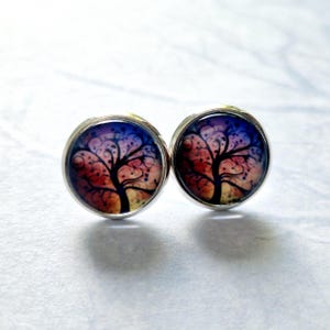 May include: A pair of silver stud earrings featuring a colourful tree design. The tree is a dark silhouette against a purple, pink and orange background.