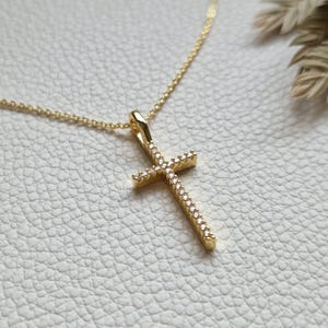 Op de afbeelding: Een goudkleurige hangerketting met een kruisontwerp. Het kruis is bezet met kleine, heldere edelstenen en hangt aan een delicate ketting. De ketting wordt gepresenteerd op een getextureerd, gebroken witte ondergrond.