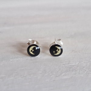 Op de afbeelding: Een paar zilveren oorstekers. Elke oorbel heeft een zwart cirkelvormig ontwerp met een crèmekleurige maan en ster. De oorbellen zijn geplaatst op een wit houten oppervlak.