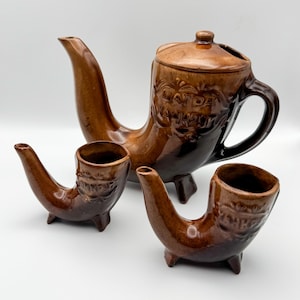 Könnte beinhalten: Ein braunes Keramik-Teeservice, bestehend aus einer Teekanne mit gebogenem Ausguss und zwei pfeifenförmigen Tassen. Die Teekanne und die Tassen haben eine glänzende Oberfläche und geprägte Details.