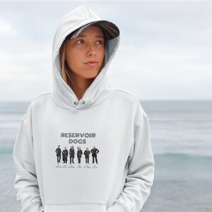 Könnte beinhalten: Ein weißer Kapuzenpullover mit einem Grafikdruck der Reservoir Dogs Filmfiguren. Der Text Reservoir Dogs steht über den Charakteren, wobei die Namen in schwarzer Schrift unter jeder Figur aufgeführt sind. Der Hoodie hat eine Vordertasche.