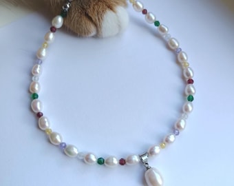 Può includere: Una delicata collana di perle con un piccolo pendente a forma di pera. La collana presenta perle bianche intervallate da perline colorate. Una zampa di gatto è visibile sullo sfondo.