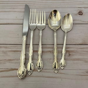 Puede incluir: Un juego de cubiertos plateados, que incluye un cuchillo, dos tenedores y dos cucharas, dispuestos sobre una superficie de madera clara. Los utensilios tienen mangos ornamentados y un acabado pulido, lo que sugiere un estilo vintage o antiguo.