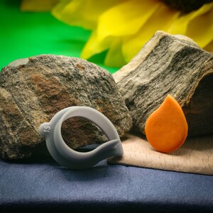 Könnte beinhalten: Ein grauer Plastikring mit der Nummer "15" darauf, ein kleines orangefarbenes tropfenförmiges Objekt und zwei graue Steine. Die Objekte befinden sich auf einer blauen Stoffoberfläche.