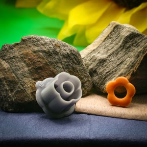 Könnte beinhalten: Zwei plastik-Blumenförmige Ausstecher, einer grau und einer orange, auf einer blauen Stoffoberfläche. Der graue Ausstecher hat die Nummer "15" an der Seite. Die Ausstecher liegen neben einem grauen Stein.