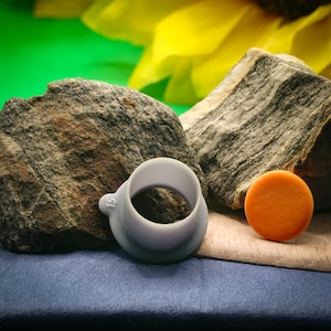 Könnte beinhalten: Ein grauer Plastikring mit der Nummer "15" darauf gedruckt. Der Ring liegt auf einer grauen Oberfläche neben einer kleinen orangen Scheibe und zwei grauen Steinen.