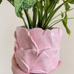 May include: A cylindrical ceramic plant pot in a glossy light pink finish, featuring a relief design of overlapping ginkgo leaves with visible veins. The pot contains a green plant with variegated leaves, showcasing a textured surface.