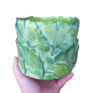Pode incluir: Um vaso de cerâmica verde com um design de folhas. O vaso cilíndrico tem uma superfície texturizada com padrões de folhas sobrepostos. O interior é de um verde mais claro. O vaso é segurado em uma mão.