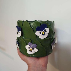 Peut inclure: Un pot de fleurs en céramique verte avec des feuilles sculptées et des accents de fleurs de pensées blanches, violettes et bleues. Le pot est cylindrique et présente une finition brillante, adapté à une utilisation intérieure ou extérieure. Le design floral ajoute une touche de décoration inspirée de la nature.
