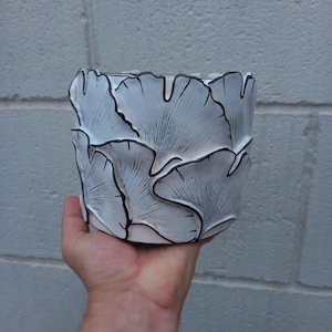 Könnte beinhalten: Weißer Keramik-Blumentopf mit Ginkgo-Blatt-Design und schwarzen Umrissen. Der zylindrische Topf hat eine strukturierte Oberfläche mit geschichteten Blättern. Das Design hat eine moderne, künstlerische Ästhetik.