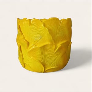 Könnte beinhalten: Ein leuchtend gelber, zylindrischer Pflanztopf mit strukturierter Oberfläche. Der Pflanztopf ist mit einem sich wiederholenden Muster stilisierter Ginkgoblätter verziert. Die Blätter sind aufwendig detailliert und verleihen dem Design Tiefe und visuelles Interesse.