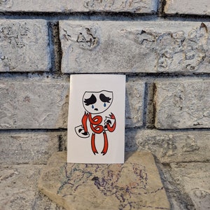 May include: A white sticker with a cartoon character wearing a red costume and holding two masks. The character has a sad expression and is crying. The character is on a white background.