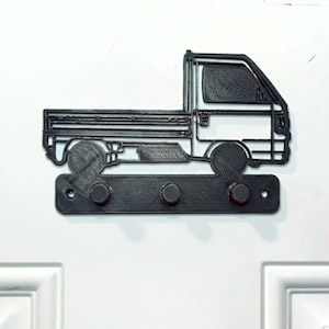 Puede incluir: Llavero negro impreso en 3D con forma de camioneta. El llavero tiene tres clavijas para colgar llaves y está montado en una puerta blanca. El diseño del camión incluye detalles de la cabina, la caja y las ruedas.