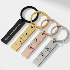 Puede incluir: Cuatro llaveros con etiquetas rectangulares y anillos redondos. Las etiquetas son negras, doradas, oro rosa y plateadas. Cada etiqueta presenta un diseño de mapa con una línea punteada y un corazón. La etiqueta negra tiene el texto "Always under the same sky".