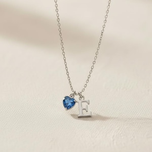 Peut inclure: Un collier en argent avec une chaîne délicate, orné d'un pendentif en forme de cœur bleu et de la lettre "E". Le pendentif en forme de cœur a un aspect facetté, et la lettre est dans une police simple sans empattement. Le collier est présenté sur un fond neutre.