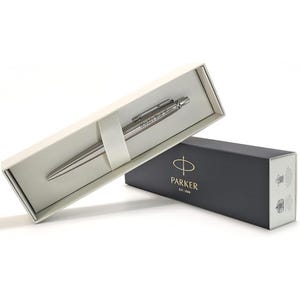 May include: A silver Parker pen, inscribed with "World's Best Mum", rests in an open cream box. A closed black box, bearing the Parker logo, sits beside it. The pen is a stylish, metallic writing instrument, ideal for gifting.