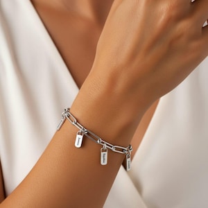 May include: A silver-colored link bracelet with several small rectangular and padlock-shaped charms. Each charm has engraved text. The bracelet is worn on a wrist, with a white garment in the background.