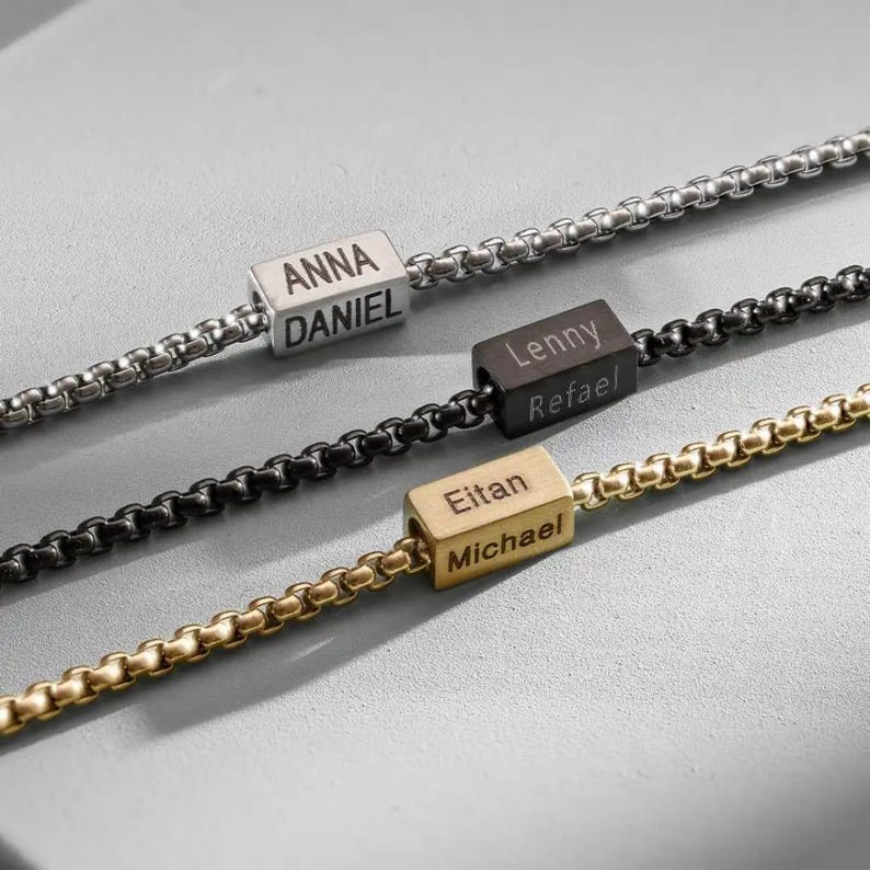 May include: Three personalized necklaces with box chains. The first is silver with "ANNA DANIEL" engraved on a silver cube. The second is black with "Lenny Refael" on a black cube. The third is gold with "Eitan Michael" on a gold cube.