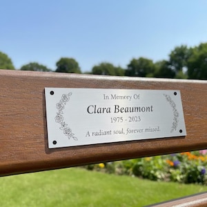 Könnte beinhalten: Rechteckige silberne Gedenktafel auf einer braunen Holzbank. Auf der Tafel steht: "In Memory Of Clara Beaumont 1975-2023 A radiant soul, forever missed." Blumendesigns umrahmen den Text. Der Hintergrund zeigt einen Park.