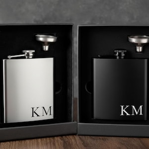 Könnte beinhalten: Zwei Flachmänner, einer silberfarben und einer schwarz, jeweils in einer schwarzen Box. Der silberne Flachmann hat die Initialen "KM" und der schwarze Flachmann hat ebenfalls die Initialen "KM". Jeder Flachmann enthält einen kleinen Trichter.