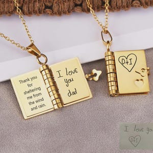 Peut inclure: Deux colliers pendentifs dorés en forme de livre. L'un est ouvert, affichant le texte gravé : "Merci de m'abriter du vent et de la pluie. Je t'aime papa." L'autre est fermé, avec un cœur et des initiales. Une petite clé est incluse.