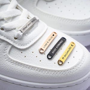 May include: White sneakers with metal shoelace tags. One tag reads "Mind over miles." Other tags are gold, black, and rose gold, with phrases "Linda & Jacob," "Stay Strong!" and "Go, Leo, GO."