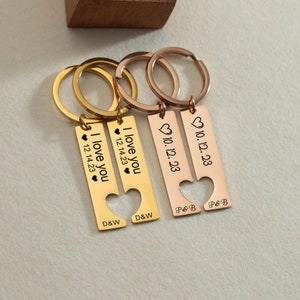 May include: Two gold and rose gold keychains with heart cutouts. The gold keychain has the text "I love you" and "12.14.23" with the initials "D&W". The rose gold keychain has the text "10.12.23" with the initials "P&B".