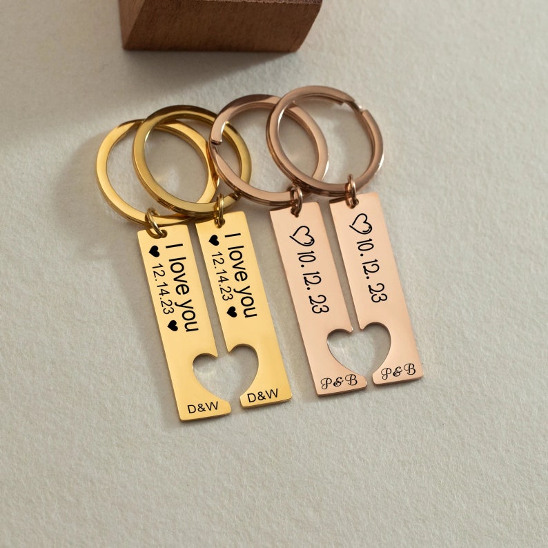 May include: Two gold and rose gold keychains with heart cutouts. The gold keychain has the text "I love you" and "12.14.23" with the initials "D&W". The rose gold keychain has the text "10.12.23" with the initials "P&B".