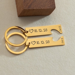 May include: Gold-toned keychain set with two matching keychains. One keychain has a heart shape cut out and the text "P&B" engraved on it. The other keychain has the text "10.12.23" and a heart symbol engraved on it.