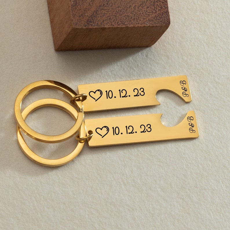 May include: Gold-toned keychain set with two matching keychains. One keychain has a heart shape cut out and the text "P&B" engraved on it. The other keychain has the text "10.12.23" and a heart symbol engraved on it.