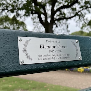 May include: A metal plaque on a green bench reads "Dedicated To Eleanor Vance 1935-2020." The plaque is adorned with floral designs and the inscription: "Her laughter brightened every day, her kindness left a lasting imprint."