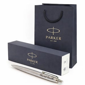 May include: A silver pen with the name "David Smith" engraved on the side, presented with a navy blue gift box and matching shopping bag. The box and bag feature the "PARKER" logo in gold.