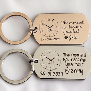 May include: Two silver keychains with a clock face and the text "The moment you became my text" with a heart and a name. The top keychain has the time 7:00 PM and the date 12-09-2021 with the name John. The bottom keychain has the time 7:00 AM and the date 20-01-2024 with the name Emily.