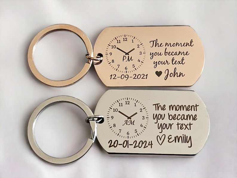 May include: Two silver keychains with a clock face and the text "The moment you became my text" with a heart and a name. The top keychain has the time 7:00 PM and the date 12-09-2021 with the name John. The bottom keychain has the time 7:00 AM and the date 20-01-2024 with the name Emily.
