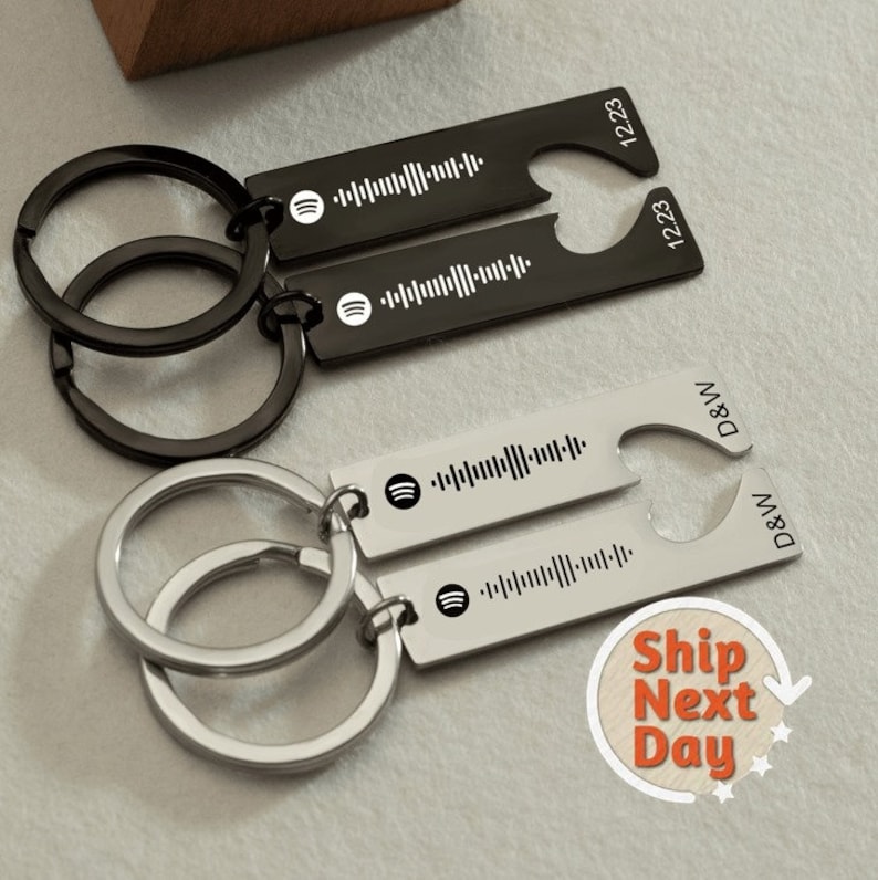May include: Two matching keychains with black and silver metal key rings. Each keychain has a silver bar with a Spotify logo and a barcode, and a heart-shaped bottle opener with the text "D&W" and "12.23".