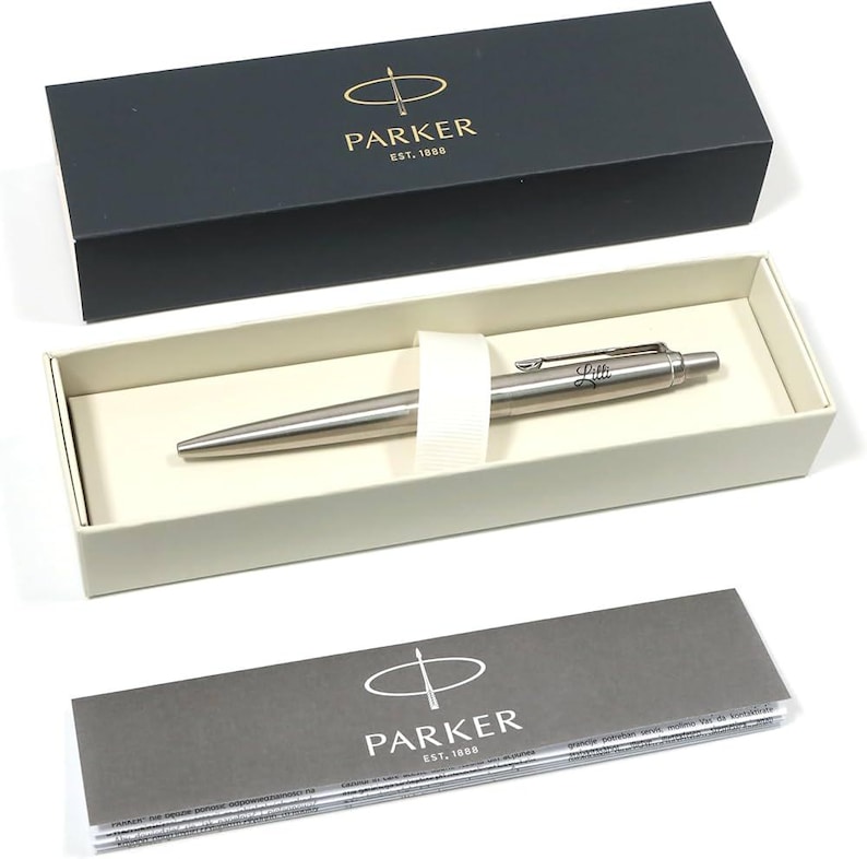May include: A silver Parker pen with the name "Lilli" engraved on the side, presented in a cream-colored box with a white ribbon. The pen is displayed inside a black box with the Parker logo and text.