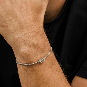 May include: A silver bracelet with a box chain design. The bracelet features a rectangular charm with the engraved names "LEO" and "ADAM". The bracelet is worn on a person's wrist, against a black background.