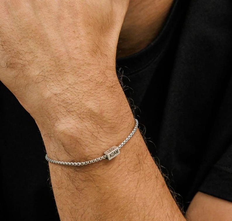May include: A silver bracelet with a box chain design. The bracelet features a rectangular charm with the engraved names "LEO" and "ADAM". The bracelet is worn on a person's wrist, against a black background.