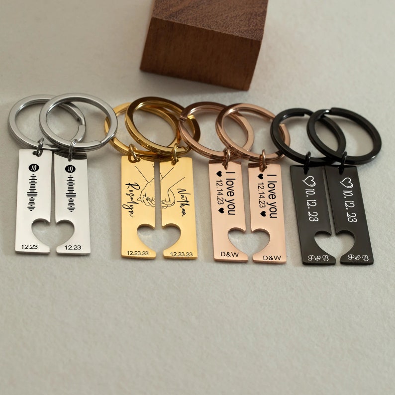 May include: Set of four matching couples keychains with heart-shaped cutouts. Each keychain features a different design, including a song lyric, a hand-holding couple, a name, and the phrase "I love you". The keychains are made of metal and come in silver, gold, rose gold, and black.