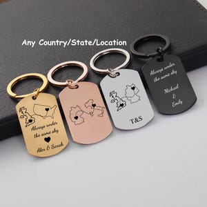 Könnte beinhalten: Vier Schlüsselanhänger aus Metall in Gold, Roségold, Silber und Schwarz. Jeder Schlüsselanhänger hat ein rechteckiges Etikett mit einem Kartendesign und dem Satz "Always under the same sky". Der Text "Any Country/State/Location" steht darüber.