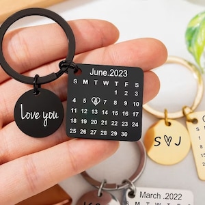 Peut inclure: Un porte-clés noir avec un charme de calendrier représentant le mois de juin 2023, un symbole de cœur le 13 et le texte "Love you" sur un charme rond. Le porte-clés comprend également un charme en or avec les initiales "SVJ" et un charme de calendrier pour octobre 2022.