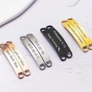 Op de afbeelding: Metalen schoenveters in goud, zilver en zwart, gegraveerd met motiverende zinnen. De tags bevatten zinnen als "Run with your heart", "Never Give Up!" en "Keep Running". Elke tag heeft een gebogen vorm met twee gaten om aan veters te bevestigen.
